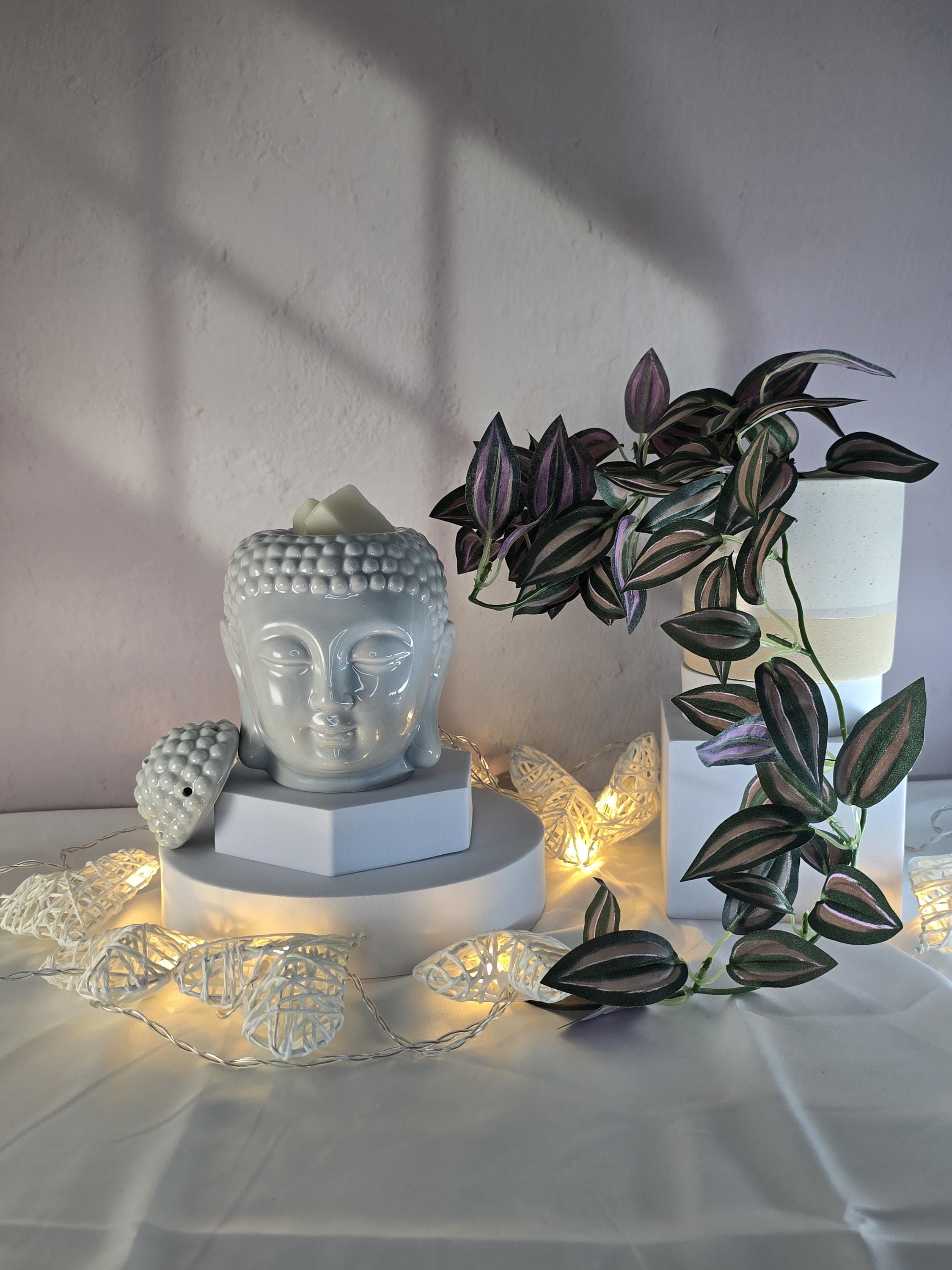Decorative setup with Buddha head sculpture, plant, and fairy lights on a white surface.
