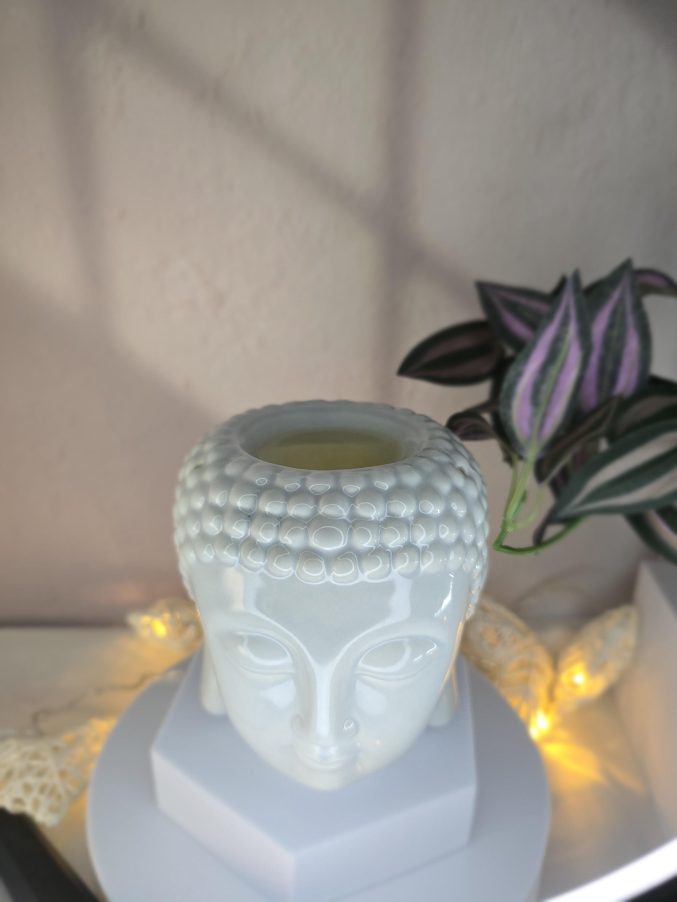White Buddha head-shaped candle holder with a lit candle inside, placed on a surface with a plant and string lights in the background.