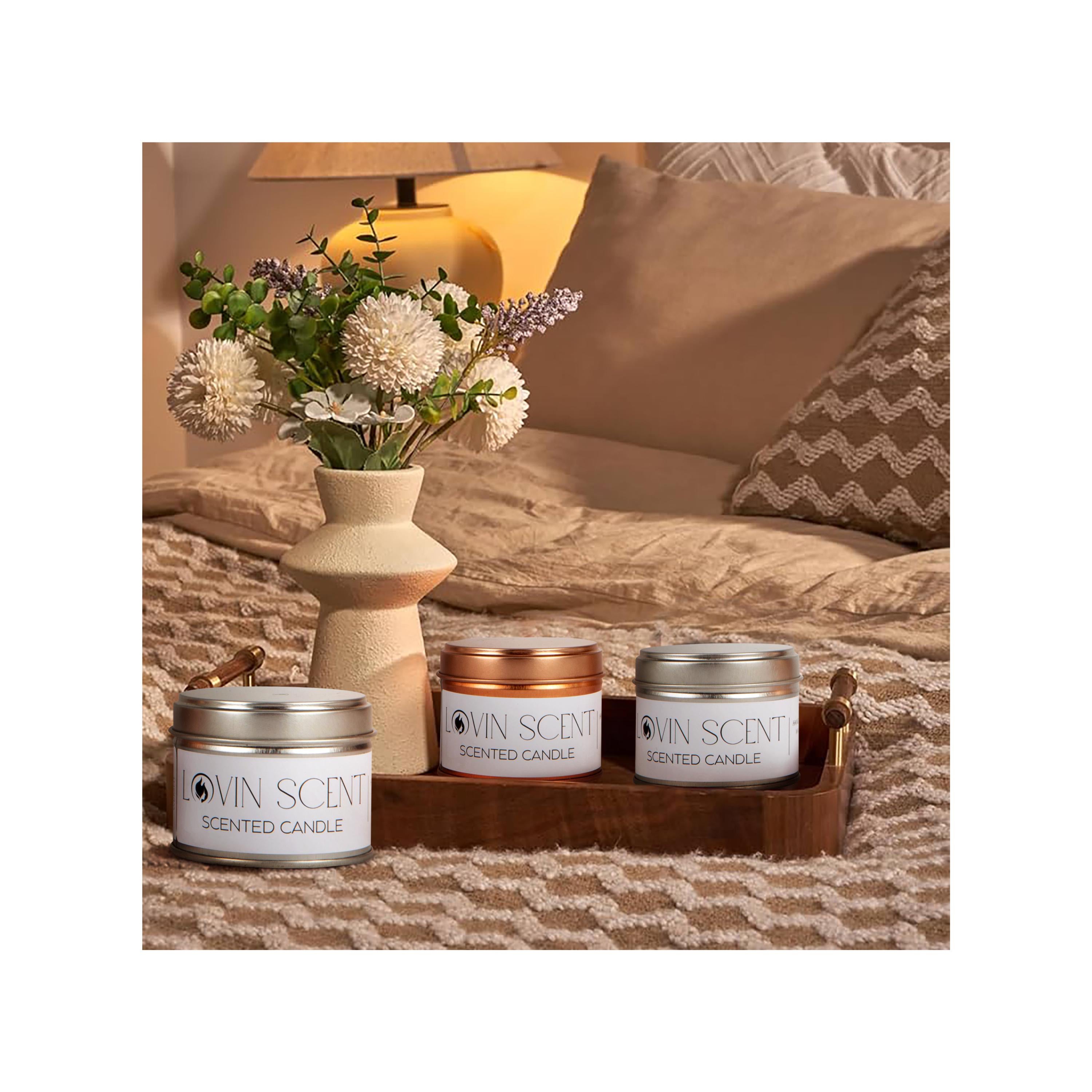 Three scented candles on a wooden tray with a vase of flowers and lamp in the background.