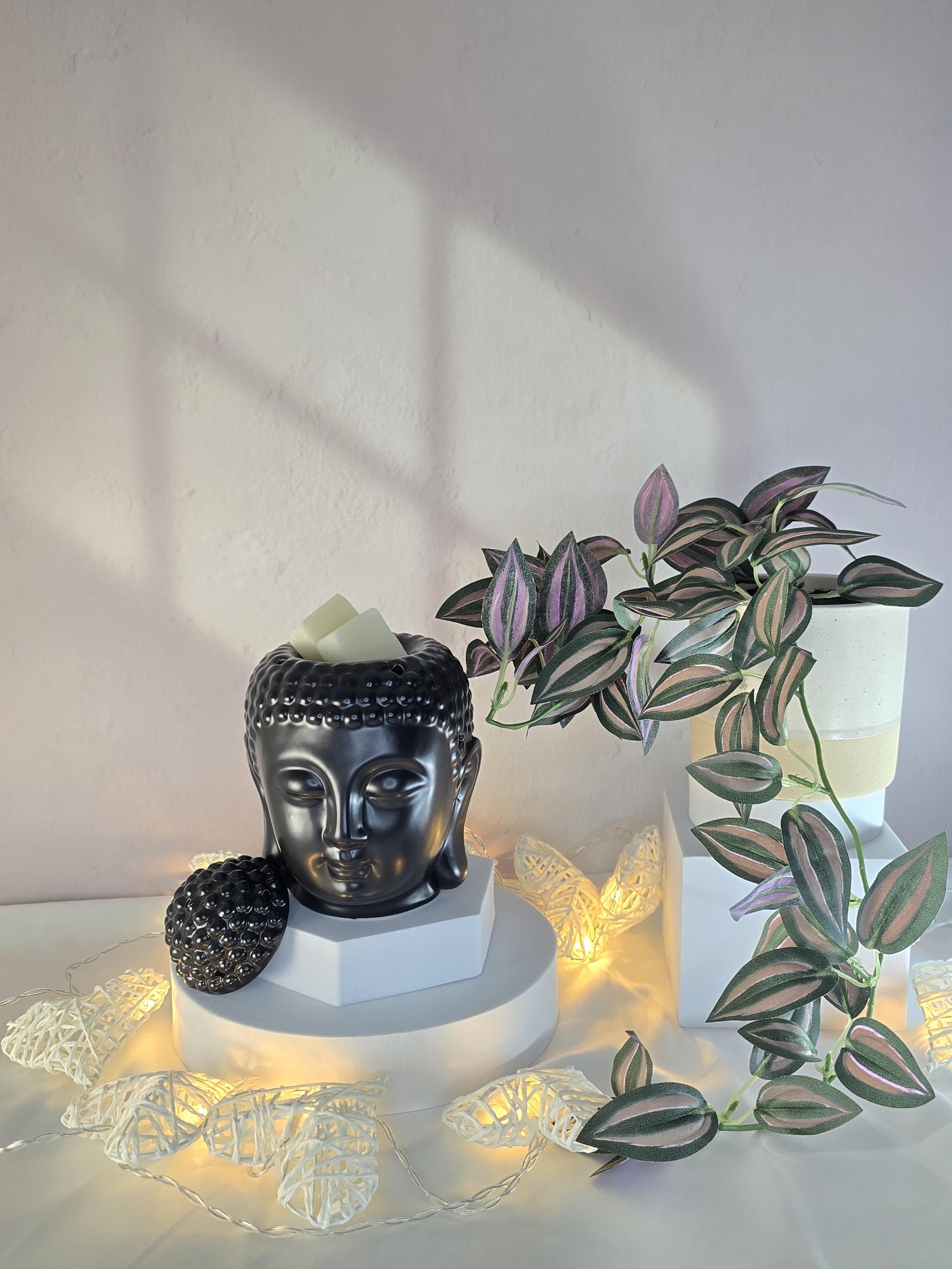 Black Buddha head sculpture with a plant and fairy lights on a white surface